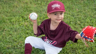 A Kyle Tee-Ball player sits on the ground holding a ball and glove with a big smile. 