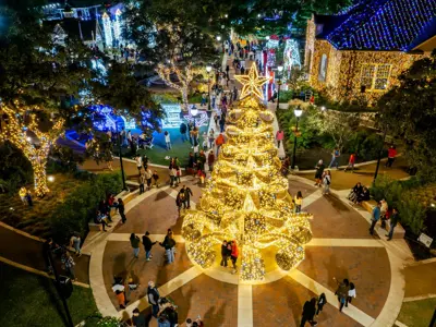A large Christmas tree adorned with golden lights stands in the center of a lively plaza at night, surrounded by people and festive buildings decorated with bright holiday lights.