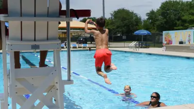 A young boy in red swim trunks jumps into the James Adkins pool while other people in the water look on. 