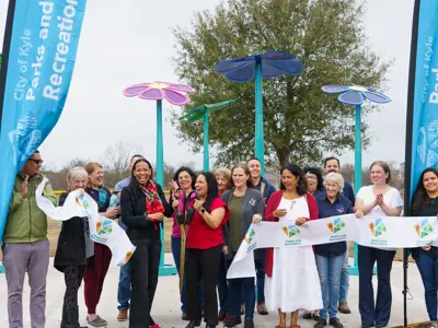 Several people stand together on a cloudy day in front of several eight to ten foot tall flower sculptures shortly after they cut a ribbon stating City of Kyle Parks and Recreation Department. 